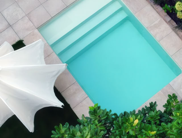 Kleiner rechteckiger Pool mit blauen Wasser, umgeben von Fliesen, einem weißen Sonnenschirm und grünen Pflanzen.