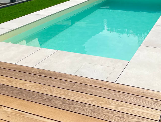 Rechteckiger Pool mit türkisfarbenem Wasser, umgeben von hellen Steinplatten, Holzterrasse im Vordergrund und Rasen im Hintergrund.