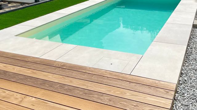 Rechteckiger Pool mit türkisfarbenem Wasser, umgeben von hellen Steinplatten, Holzterrasse im Vordergrund und Rasen im Hintergrund.