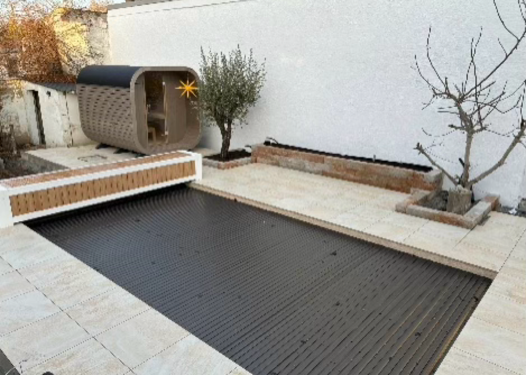 Ein Garten mit einem rechteckigen Pool, der mit einer Abdeckung versehen ist, einer kleinen Holzsauna, einem Baum und einem Olivenbaum in Töpfen.